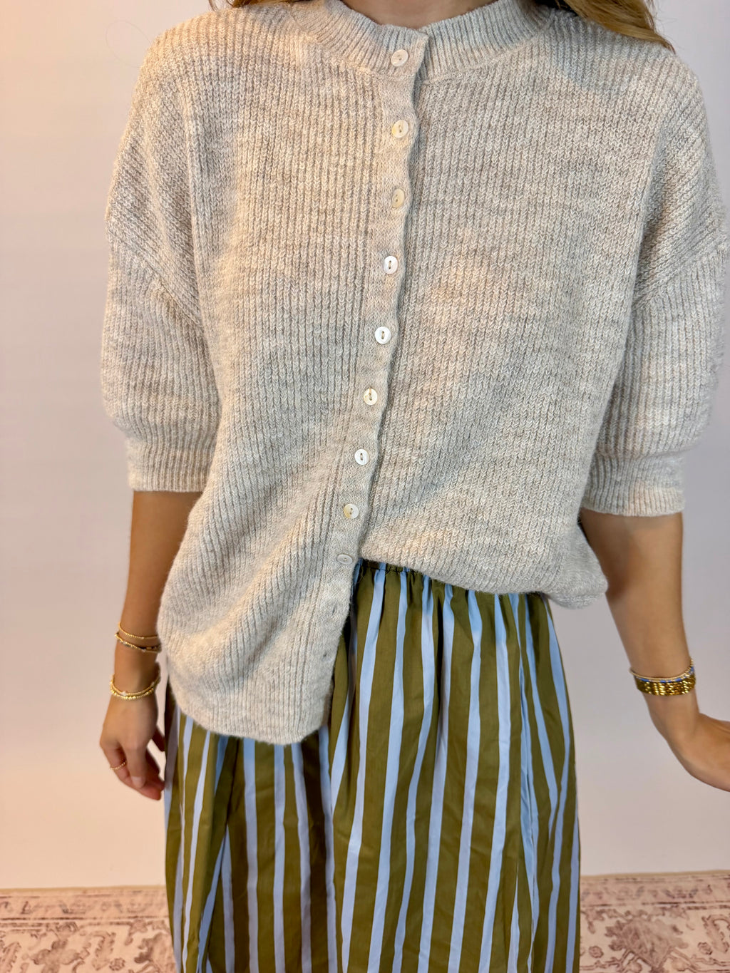 A person wearing a light oatmeal heathered ribbed knit cardigan with white buttons and olive green and white vertically striped pants, standing against a plain light wall.
