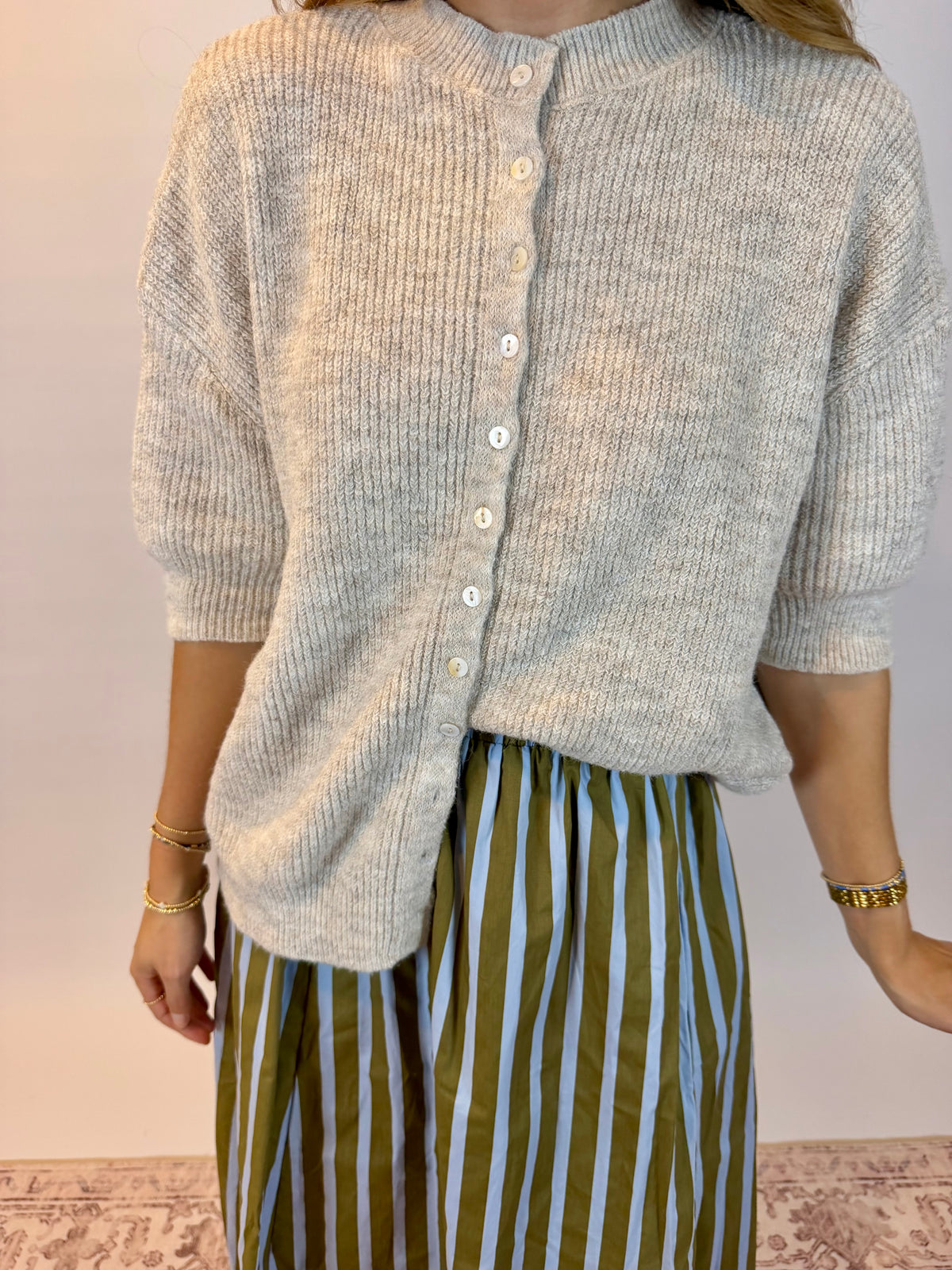 A person wearing a light oatmeal heathered ribbed knit cardigan with white buttons and olive green and white vertically striped pants, standing against a plain light wall.