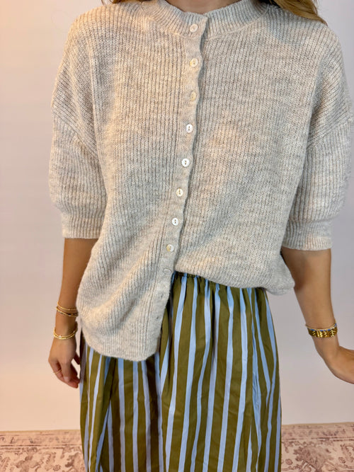 A person wearing a light oatmeal heathered ribbed knit cardigan with white buttons and olive green and white vertically striped pants, standing against a plain light wall.