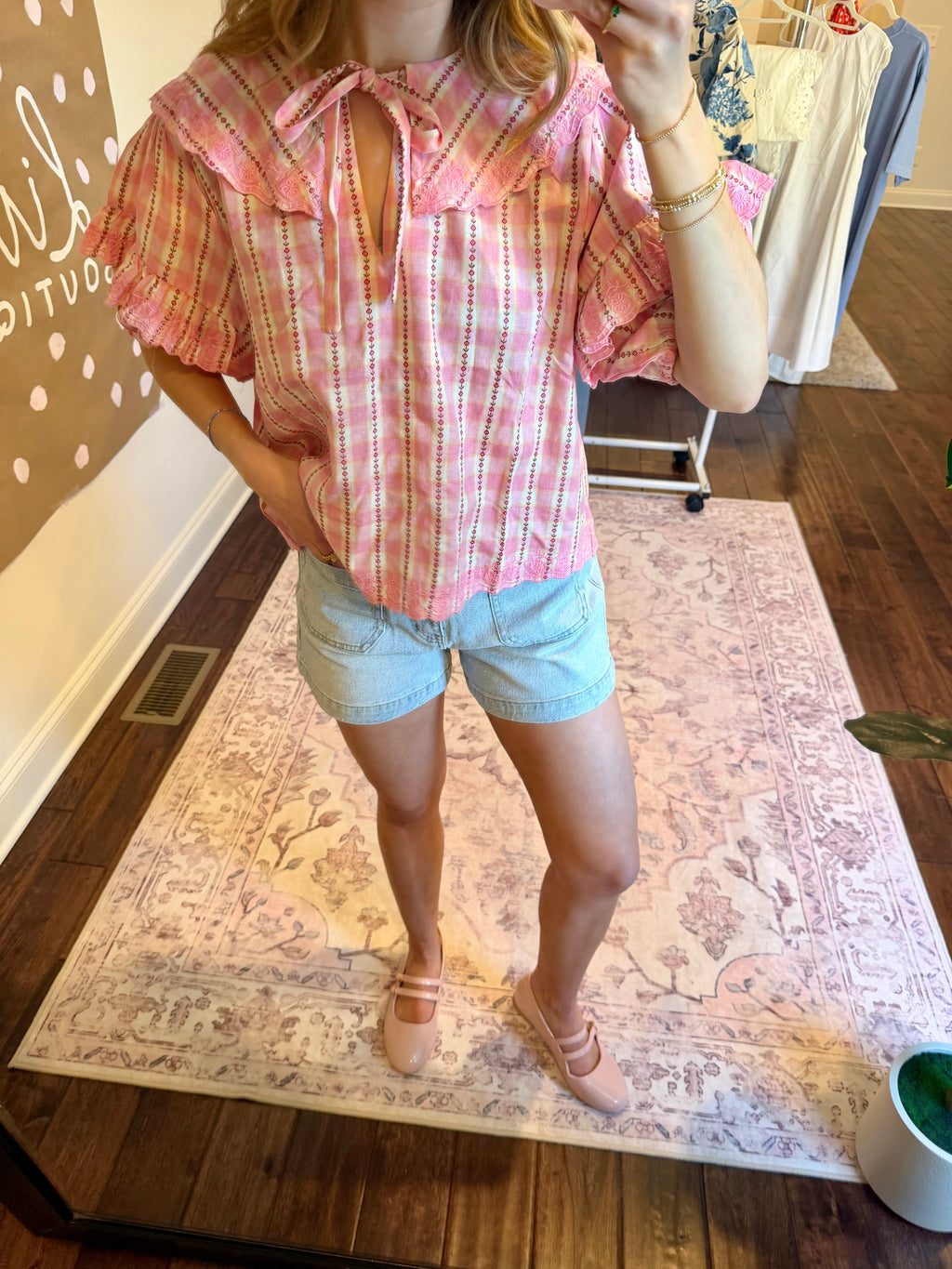 Woman taking a mirror selfie wearing a pink plaid shirt and denim shorts in a room with a chandelier and clothing rack.