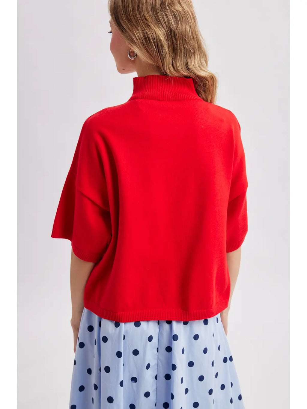 A woman shown from the back wearing a bright red short-sleeve mock neck knit top and a light blue skirt with dark blue polka dots, against a light gray studio background.