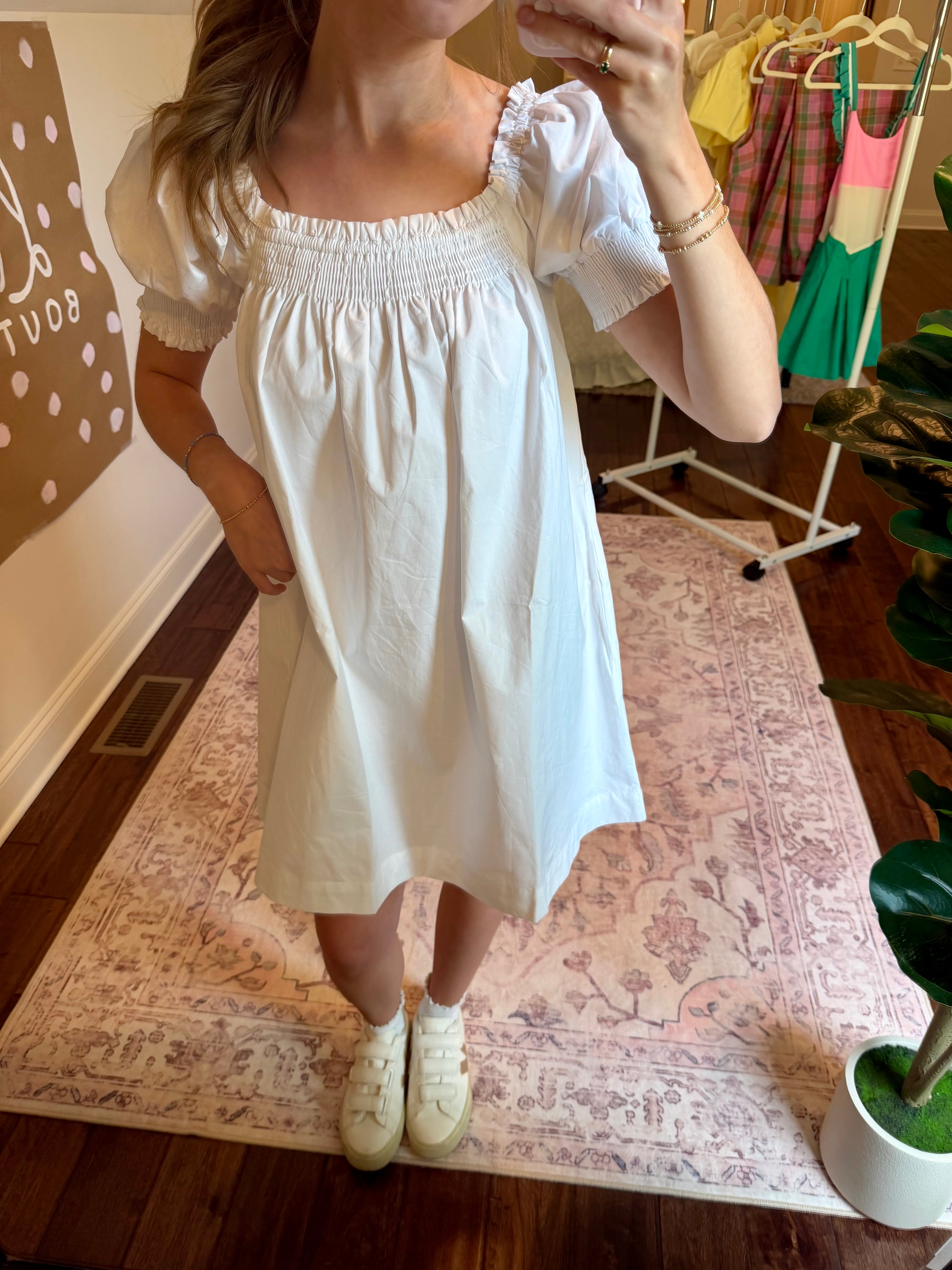 Person wearing a white dress standing on a patterned rug in a room with clothing racks.