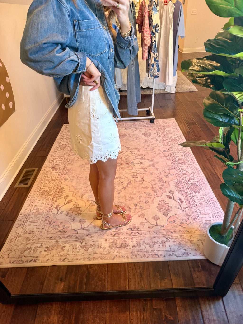 Person taking a mirror selfie wearing a denim jacket and white lace skirt in a room with a rug and plant.