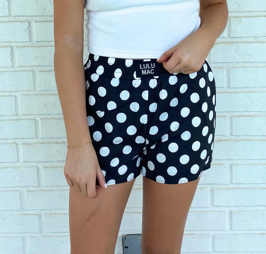 A person wearing black shorts with a white polka dot pattern, paired with a white tank top, standing against a white painted brick wall. The shorts feature a white elastic waistband with the text 'LULU MAC' printed on it.