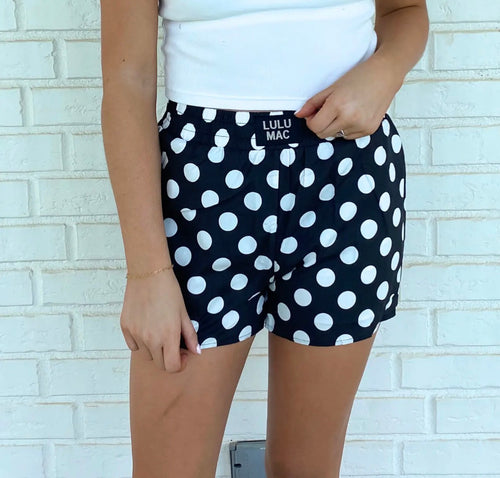 A person wearing black shorts with a white polka dot pattern, paired with a white tank top, standing against a white painted brick wall. The shorts feature a white elastic waistband with the text 'LULU MAC' printed on it.