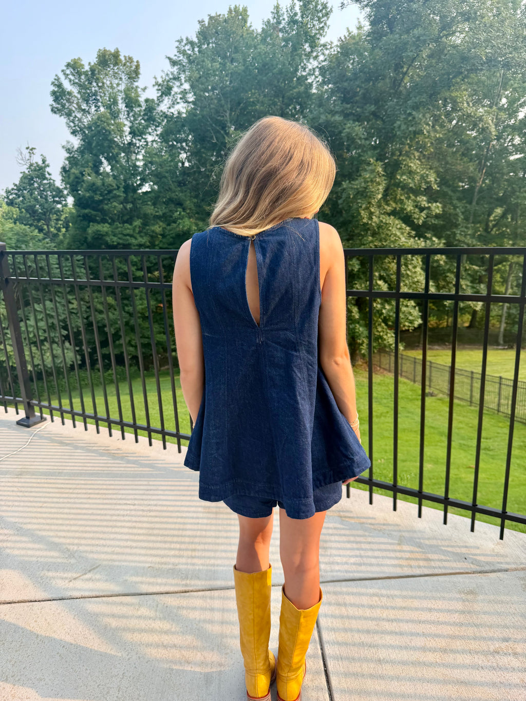 A woman stands on a patio from behind wearing a sleeveless navy blue denim dress with a back keyhole cutout and yellow rain boots, with a black railing and green trees in the background.