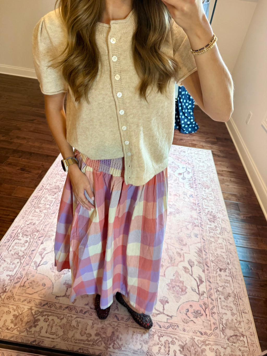 A woman wearing a cream short-sleeve button-front cardigan and a pink, purple, and white plaid maxi skirt, standing on a patterned rug in a home setting.