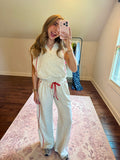 A woman takes a mirror selfie in a room with wood floors and a pink patterned rug, wearing a light beige two-piece loungewear set consisting of a sleeveless quarter-zip top and matching wide-leg sweatpants with red drawstrings and side stripes.