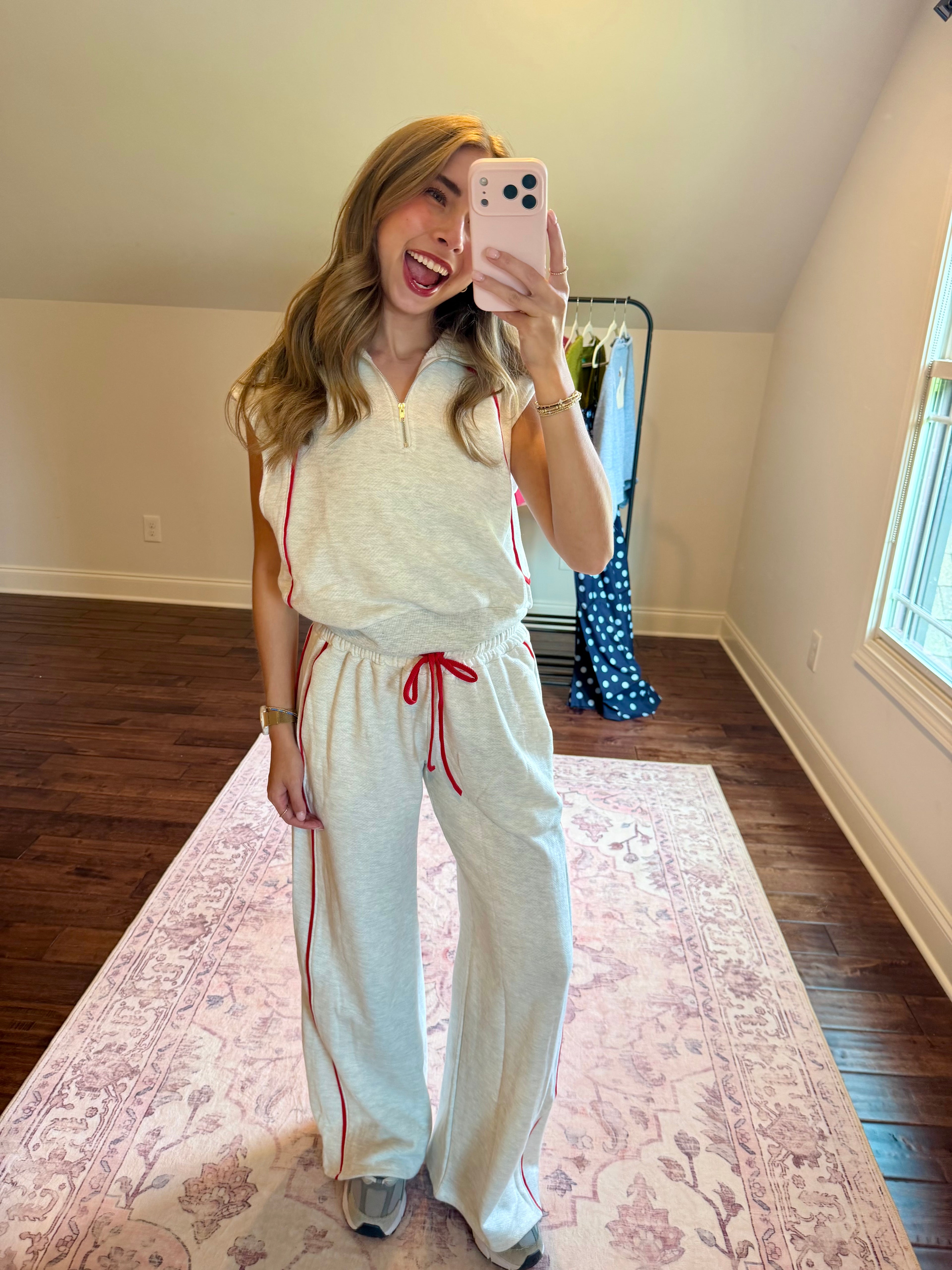 A woman takes a mirror selfie in a room with wood floors and a pink patterned rug, wearing a light beige two-piece loungewear set consisting of a sleeveless quarter-zip top and matching wide-leg sweatpants with red drawstrings and side stripes.
