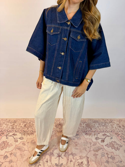 A woman wearing a dark blue cropped denim jacket with wide three-quarter sleeves, cream linen pants, and white platform sandals with black stripes, standing on a patterned pink rug against a plain light wall.