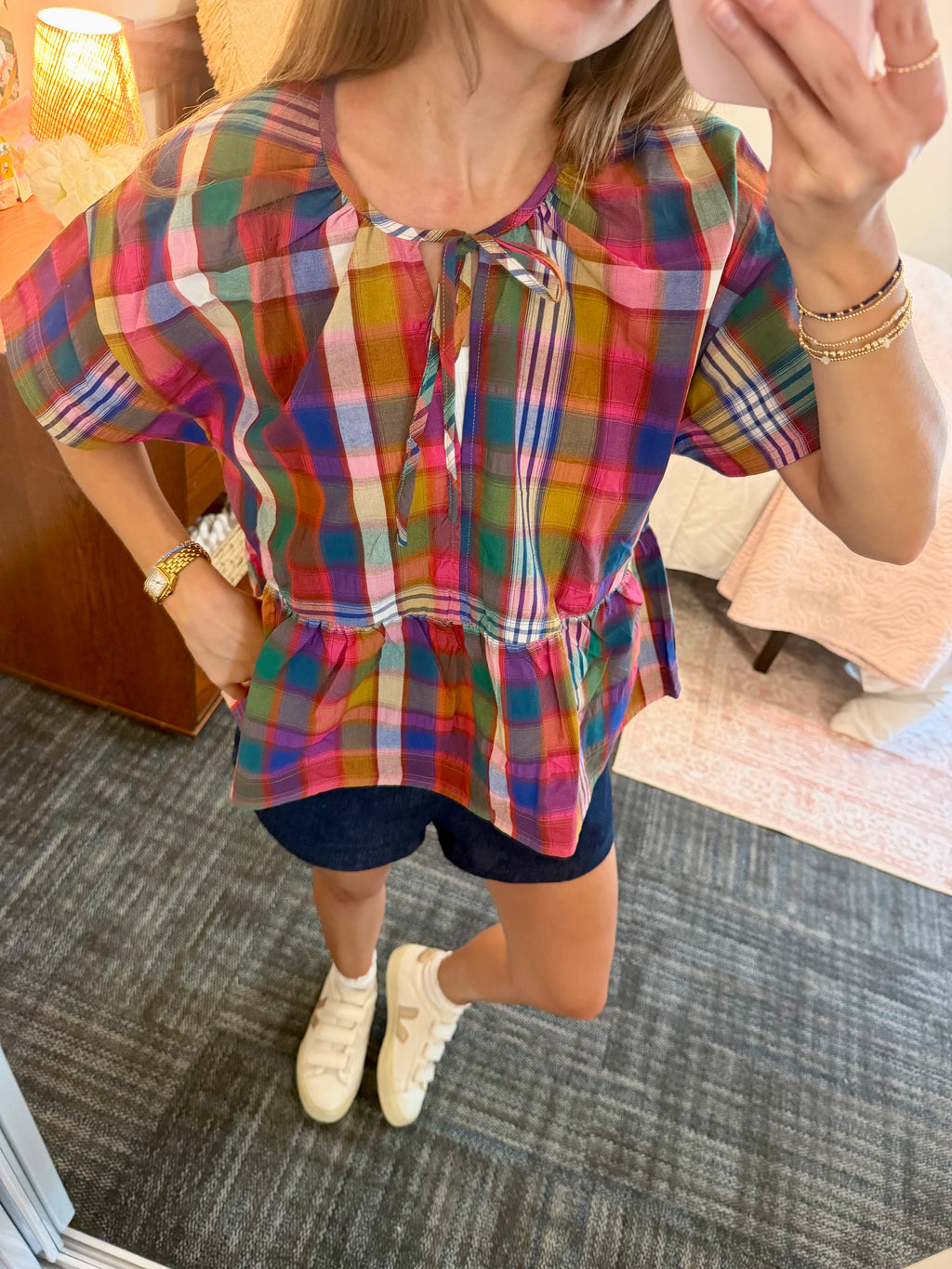 A woman wearing a colorful plaid short-sleeve blouse with a tie neckline and peplum hem, paired with dark shorts and white sneakers, taking a mirror selfie in a bedroom setting.