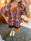 A woman wearing a colorful plaid short-sleeve blouse with a tie neckline and peplum hem, paired with dark shorts and white sneakers, taking a mirror selfie in a bedroom setting.