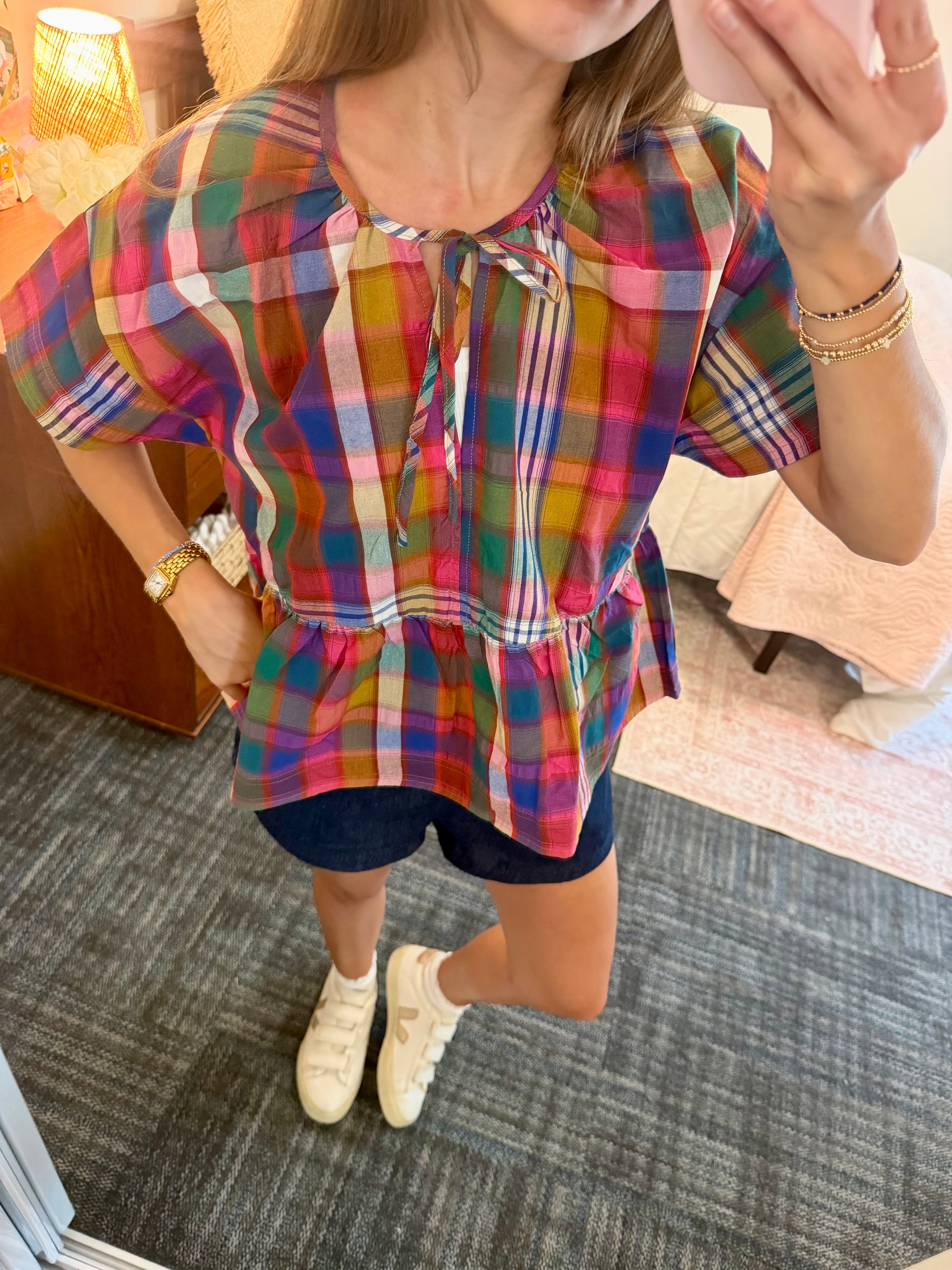 A woman wearing a colorful plaid short-sleeve blouse with a tie neckline and peplum hem, paired with dark shorts and white sneakers, taking a mirror selfie in a bedroom setting.