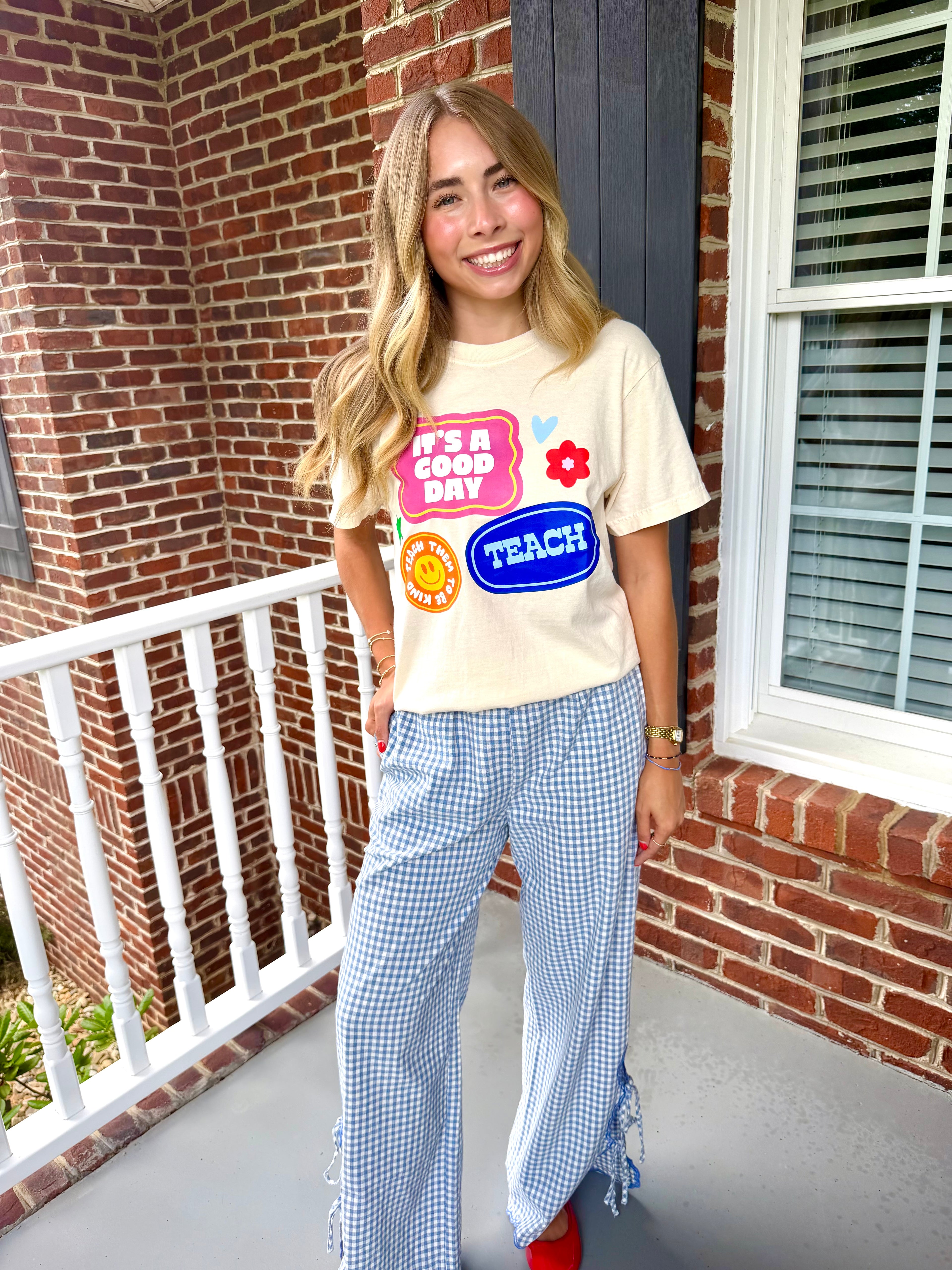A woman stands on a brick porch wearing a cream graphic T-shirt with colorful retro-style patches and blue-and-white gingham wide-leg pants with ruffle details at the hems.