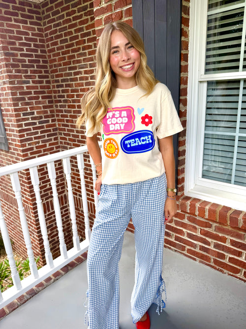 A woman stands on a brick porch wearing a cream graphic T-shirt with colorful retro-style patches and blue-and-white gingham wide-leg pants with ruffle details at the hems.