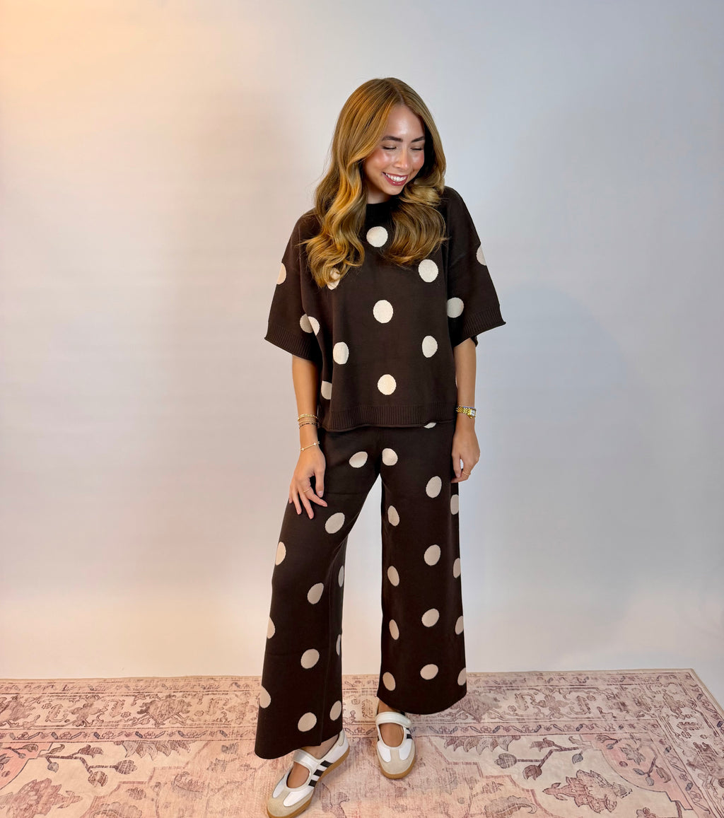A woman modeling a matching dark brown two-piece set with large white polka dots, consisting of a short-sleeve top and wide-leg pants, standing on a patterned rug against a plain light gray wall.
