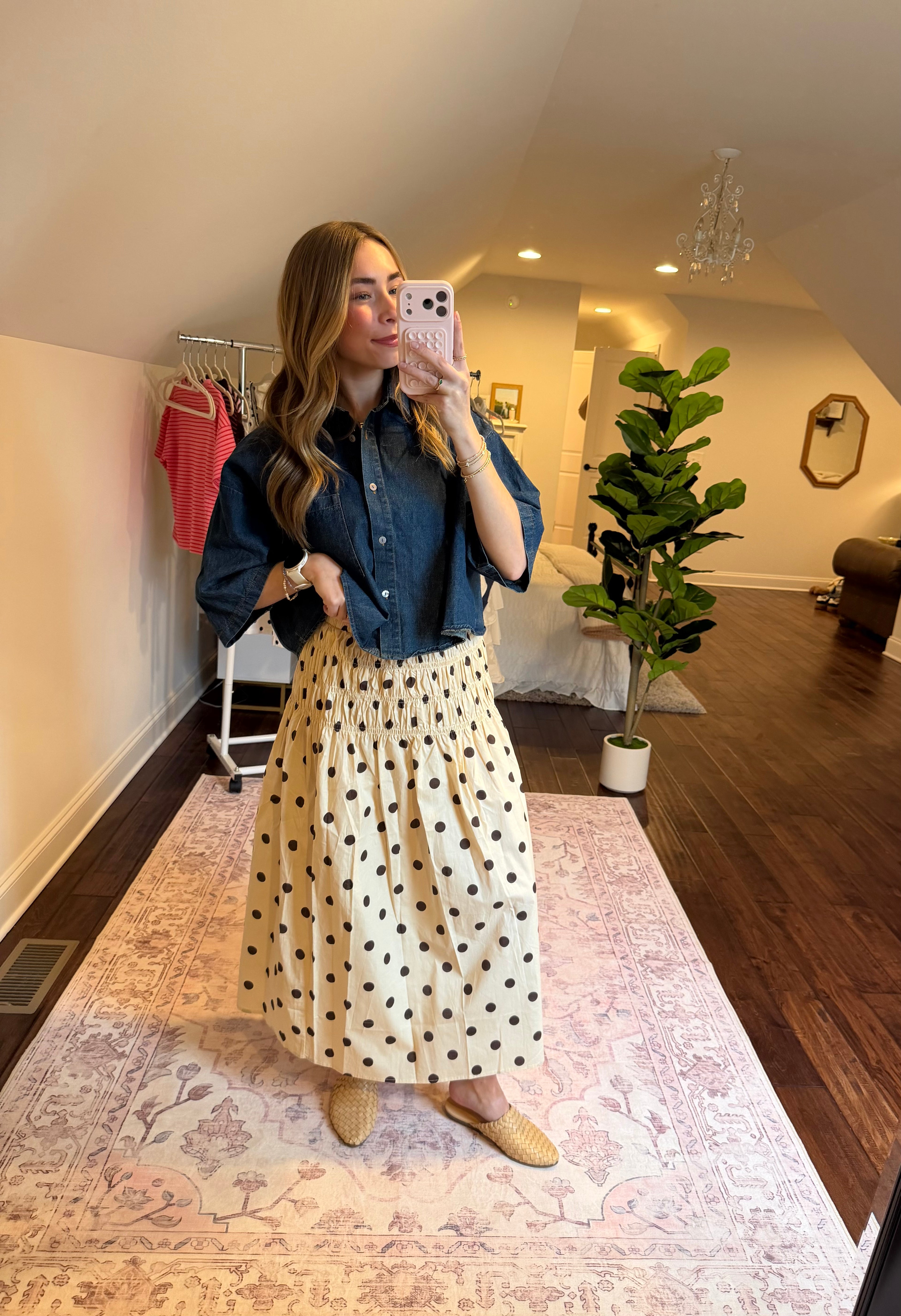 Woman taking a mirror selfie wearing a denim shirt and polka dot skirt in a room with a plant and coat rack.