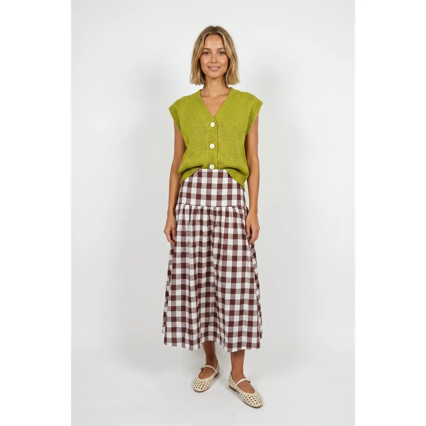 A woman modeling a sleeveless lime green knit vest with white buttons and a brown and white gingham midi skirt, paired with beige woven flat shoes, against a light gray studio background.