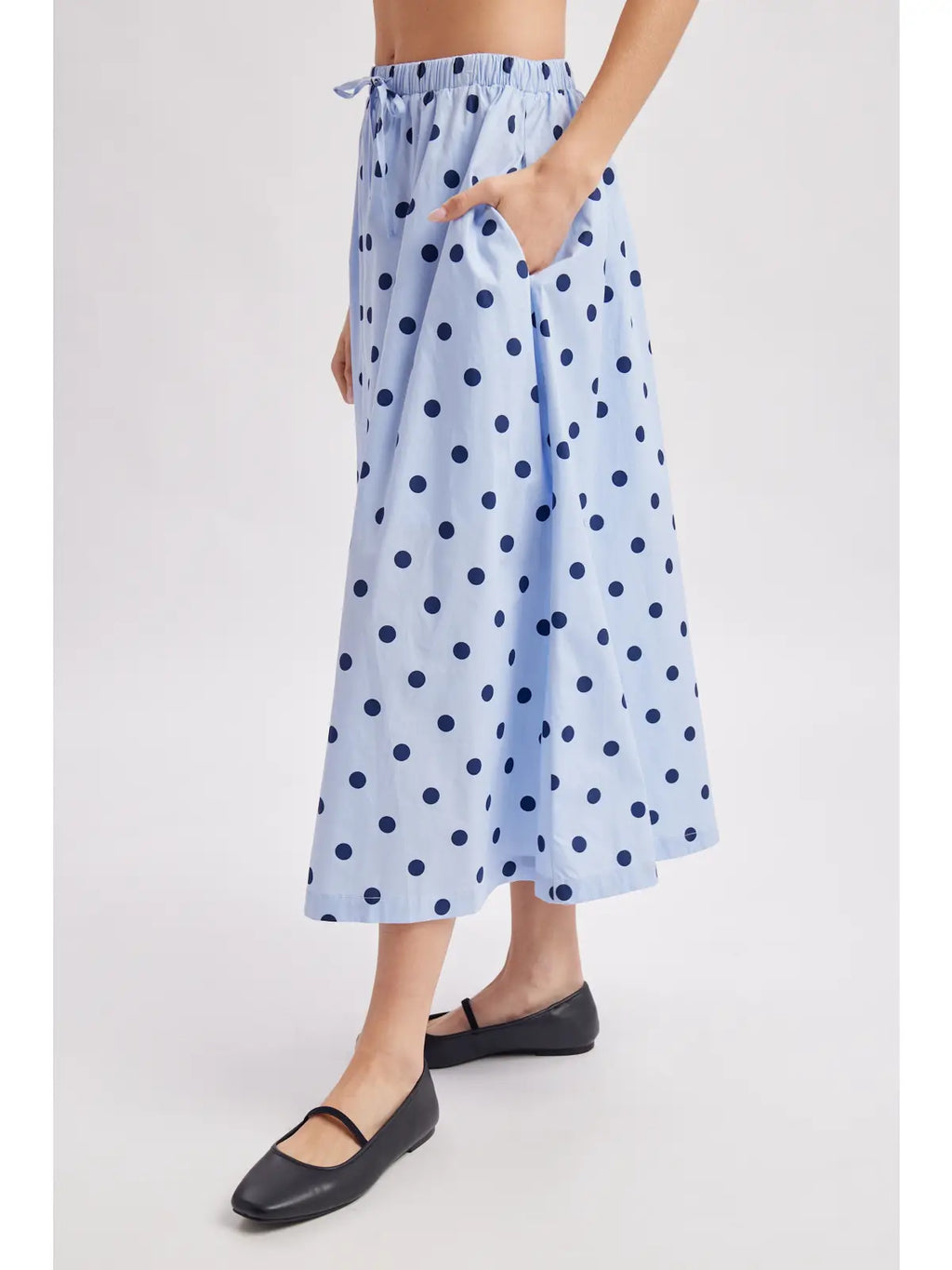 A model wearing a light blue midi skirt with dark blue polka dots, featuring an elastic waistband and side pockets, paired with black flats against a light gray background.