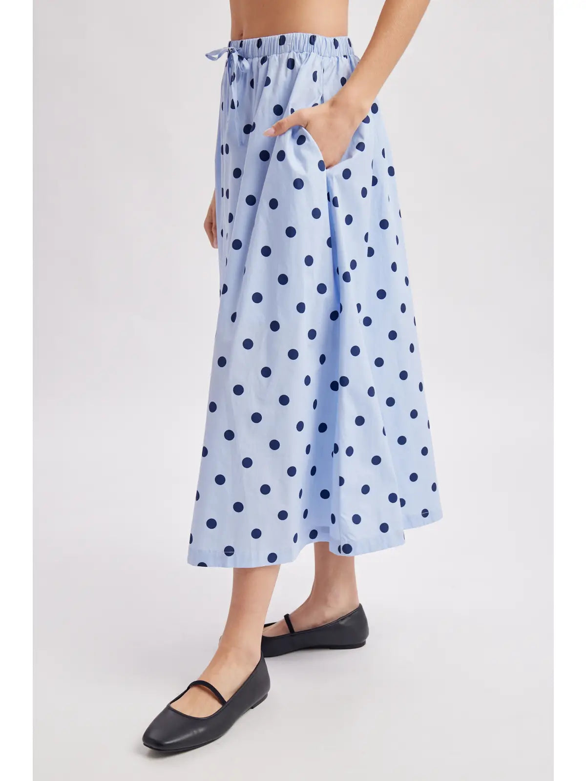 A model wearing a light blue midi skirt with dark blue polka dots, featuring an elastic waistband and side pockets, paired with black flats against a light gray background.