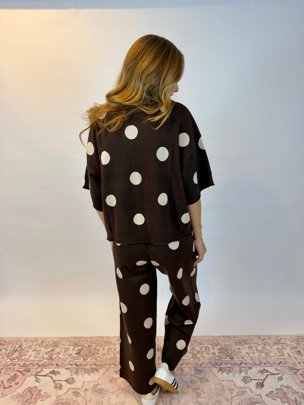 A woman is shown from behind wearing a matching dark brown knit lounge set with large white polka dots, consisting of a short-sleeve top and wide-leg pants, standing on a patterned rug against a plain wall.