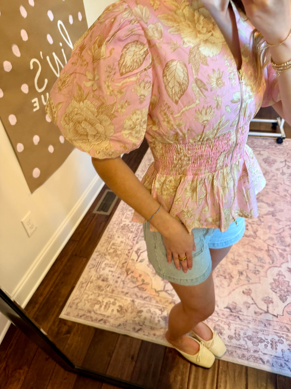 Woman taking a mirror selfie wearing a floral top and denim shorts in a home setting.