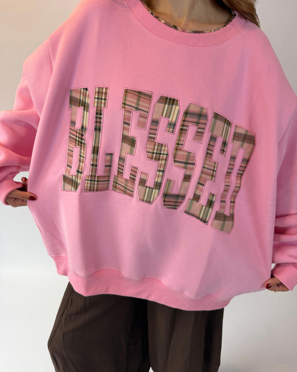 A person wearing an oversized pink sweatshirt with the word 'BLESSED' in large plaid-patterned letters across the front, paired with dark brown pants.