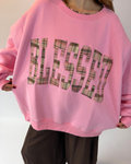 A person wearing an oversized pink sweatshirt with the word 'BLESSED' in large plaid-patterned letters across the front, paired with dark brown pants.