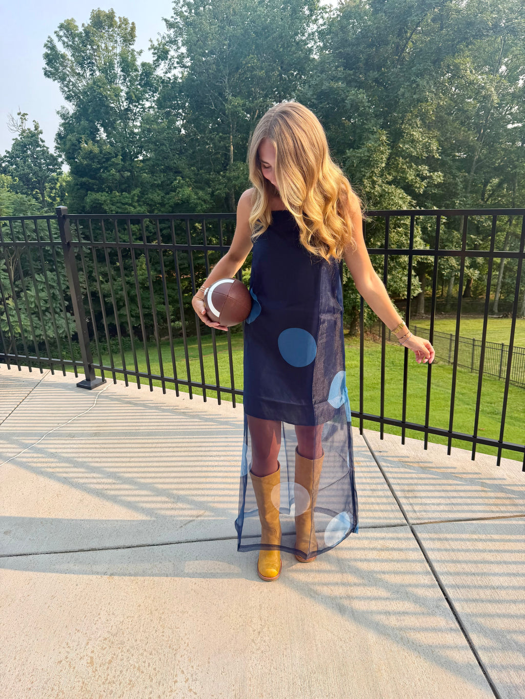 A woman stands on an outdoor patio wearing a sleeveless navy dress with large blue polka dots and a sheer overlay, paired with tan boots, while holding a football. Trees and a black metal fence are visible in the background.