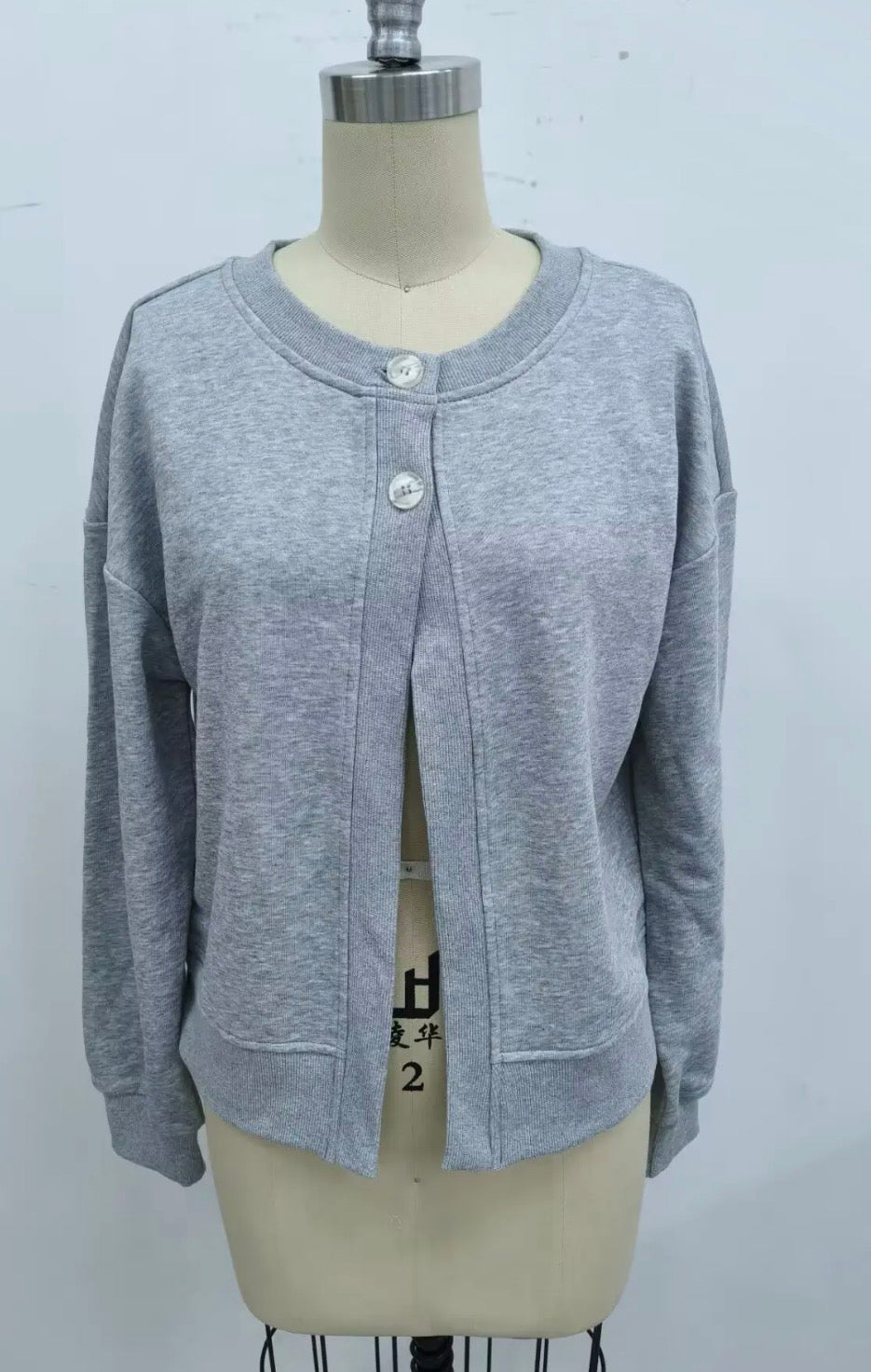 A light gray heathered knit cardigan sweater displayed on a beige dress form mannequin. The cardigan features a crew neck, three silver buttons, and a front split opening.