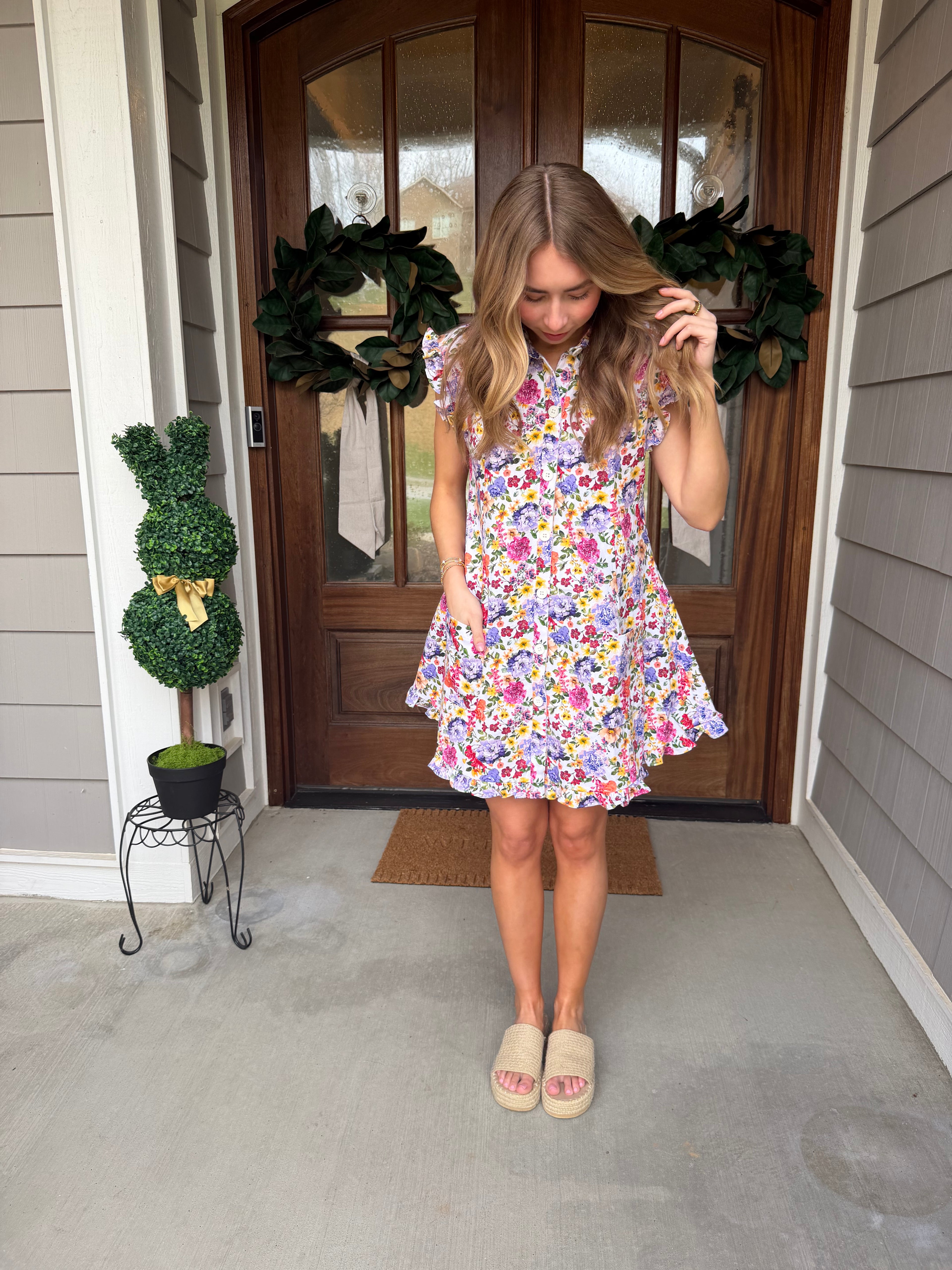 Woman in a floral dress standing in front of a wooden door with plants around.