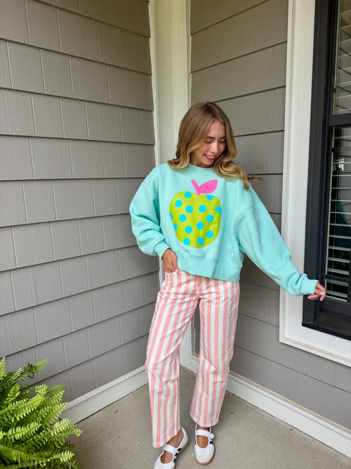 A woman stands on a porch wearing a light aqua sweatshirt with a large yellow and blue polka-dot apple graphic on the front, paired with pink and white vertically striped pants and white shoes.