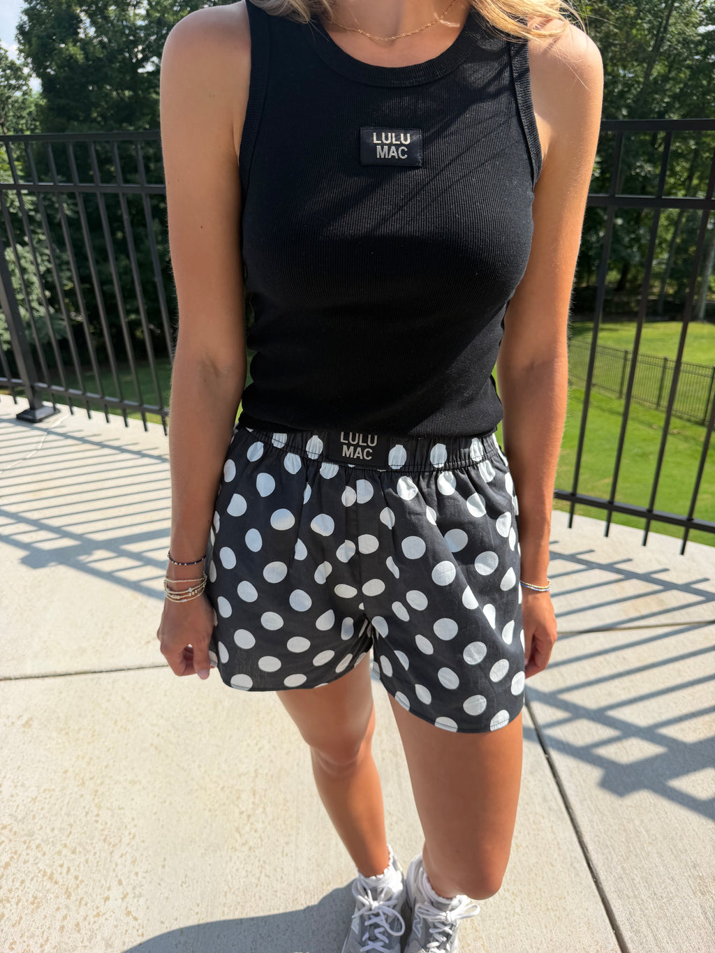 A woman wearing a black sleeveless tank top and black shorts with white polka dots, standing outdoors on a concrete patio with a black metal fence and green grass in the background.