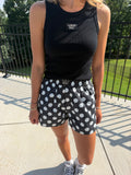 A woman wearing a black sleeveless tank top and black shorts with white polka dots, standing outdoors on a concrete patio with a black metal fence and green grass in the background.