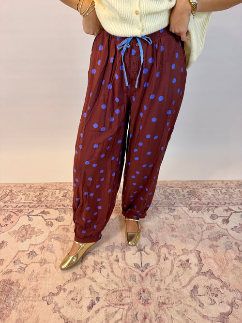 A person wearing loose brown harem-style pants with blue polka dots and a blue drawstring, paired with a cream ribbed top and metallic gold shoes, standing on a patterned rug against a plain wall.
