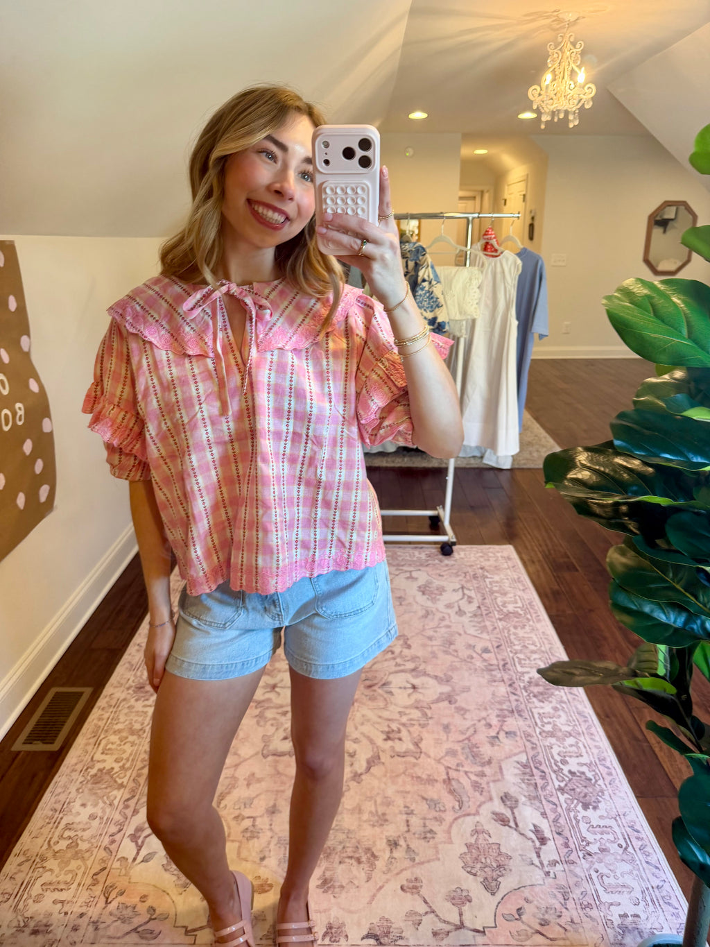 Woman taking a mirror selfie wearing a pink plaid shirt and denim shorts in a room with a chandelier and clothing rack.