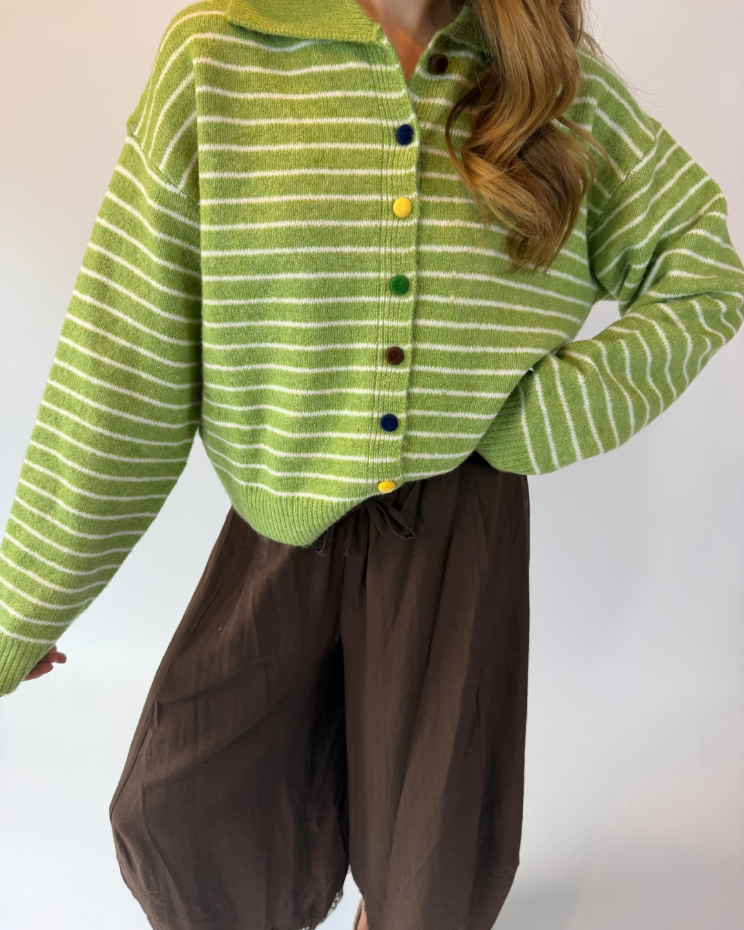 A person wearing a lime green and white striped knit cardigan with colorful buttons, paired with dark brown pants, against a plain light background.
