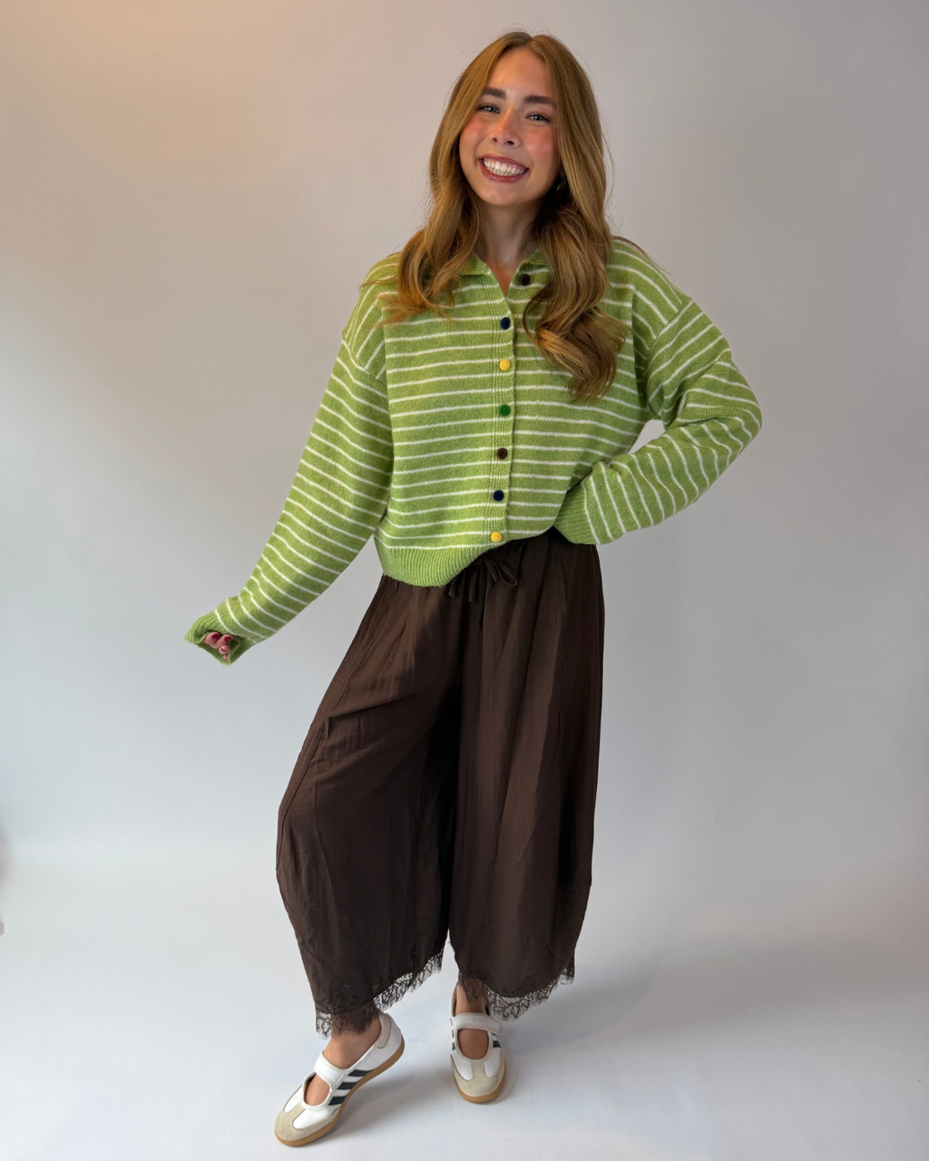 A woman modeling a light green and white striped cropped button-up cardigan, paired with brown wide-leg pants and white slide sandals, against a plain light gray studio background.