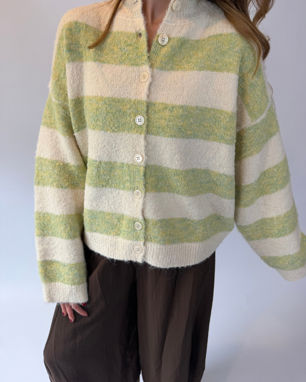 A person wearing a fuzzy knit cardigan with horizontal light green and cream stripes, paired with dark brown pants, against a plain light gray background.