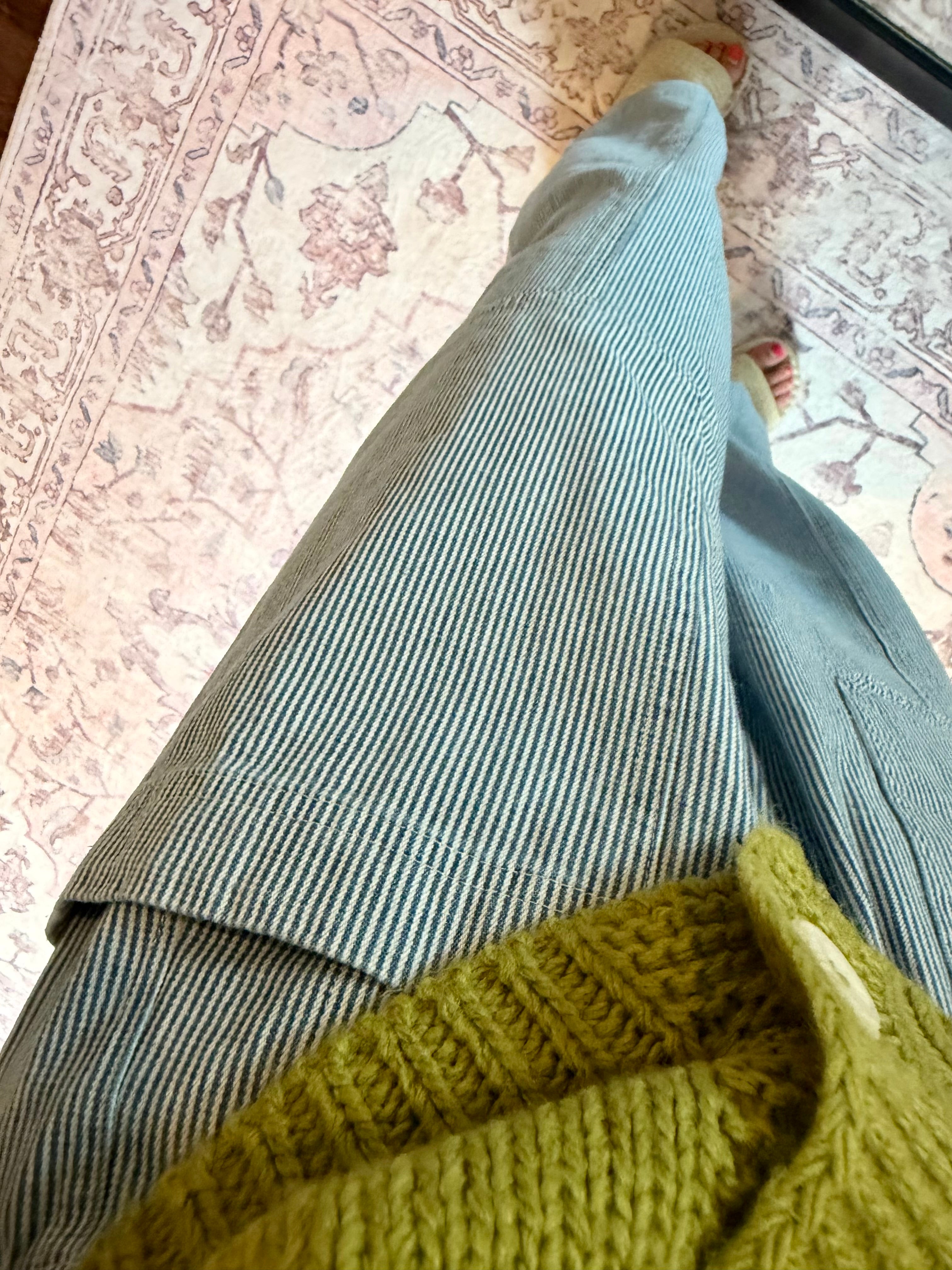 A close-up, high-angle view of a person wearing wide-leg, light blue-gray striped trousers and a chunky olive green knit sweater, standing on a patterned pink and beige rug.