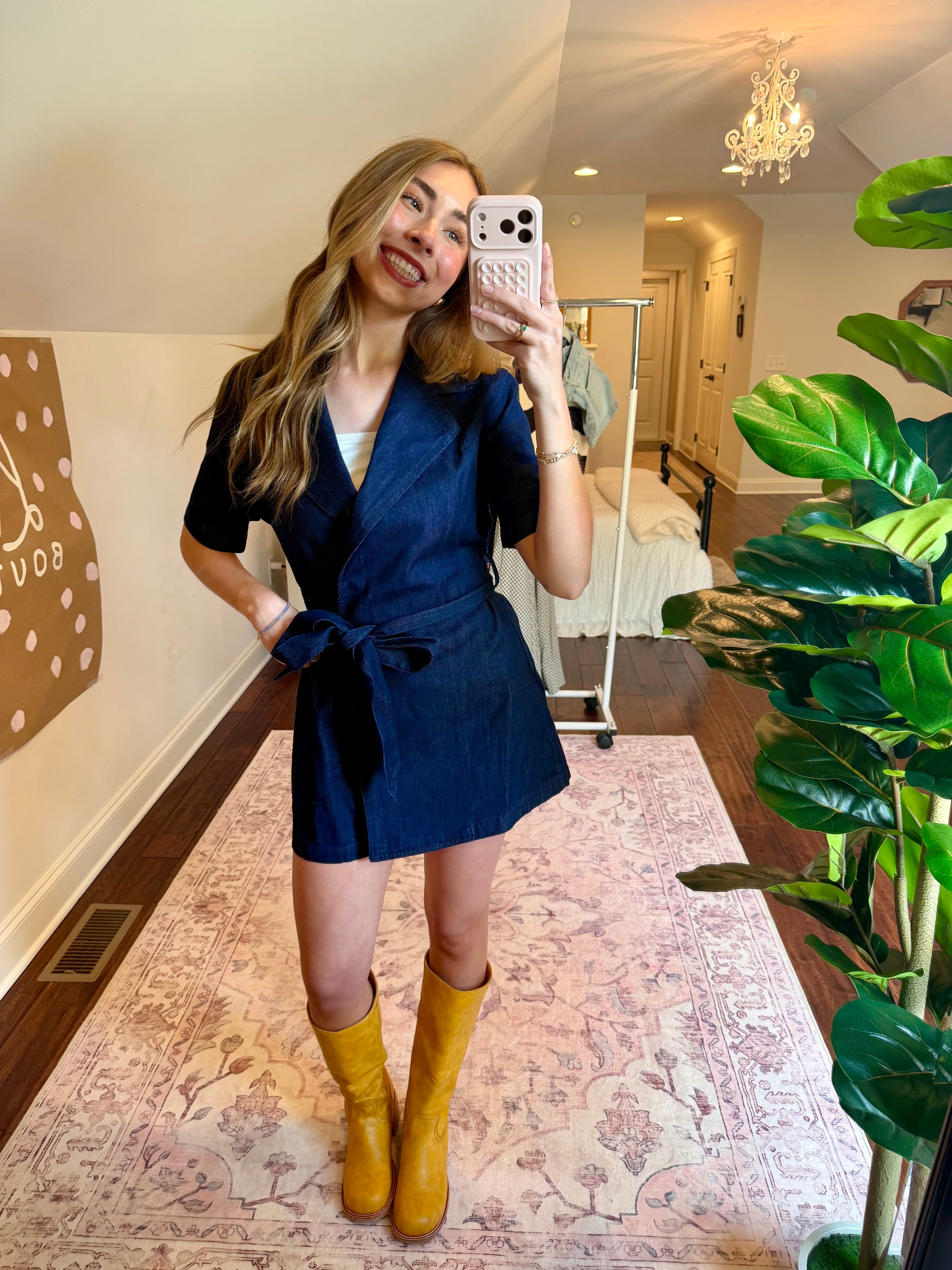 Woman taking a mirror selfie wearing a blue dress and yellow boots in a room with a plant and decorative wall art.