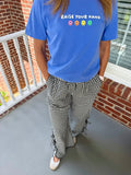 A person standing on a porch wearing a bright blue graphic T-shirt with the text 'RAISE YOUR HAND' and smiley faces, paired with black-and-white gingham wide-leg pants with ruffled trim and white sneakers.