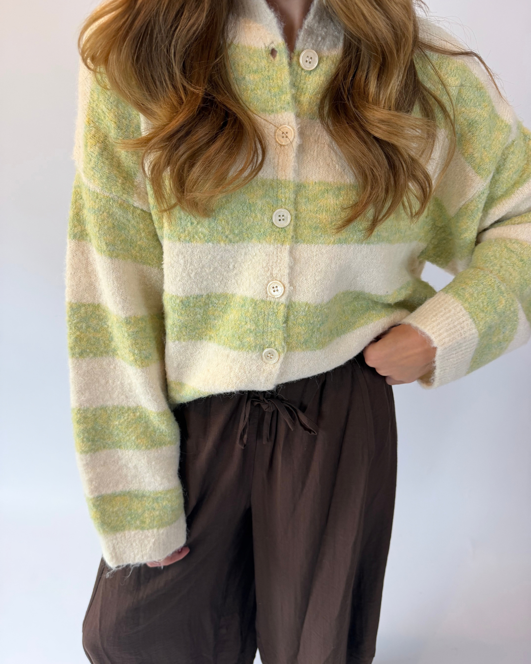 A person wearing a fuzzy knit cardigan with horizontal light green and cream stripes, paired with dark brown drawstring pants, against a plain light background.
