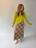 A woman modeling a bright yellow knit sweater and a brown and beige plaid midi skirt with white sneakers, standing against a plain light gray studio background.