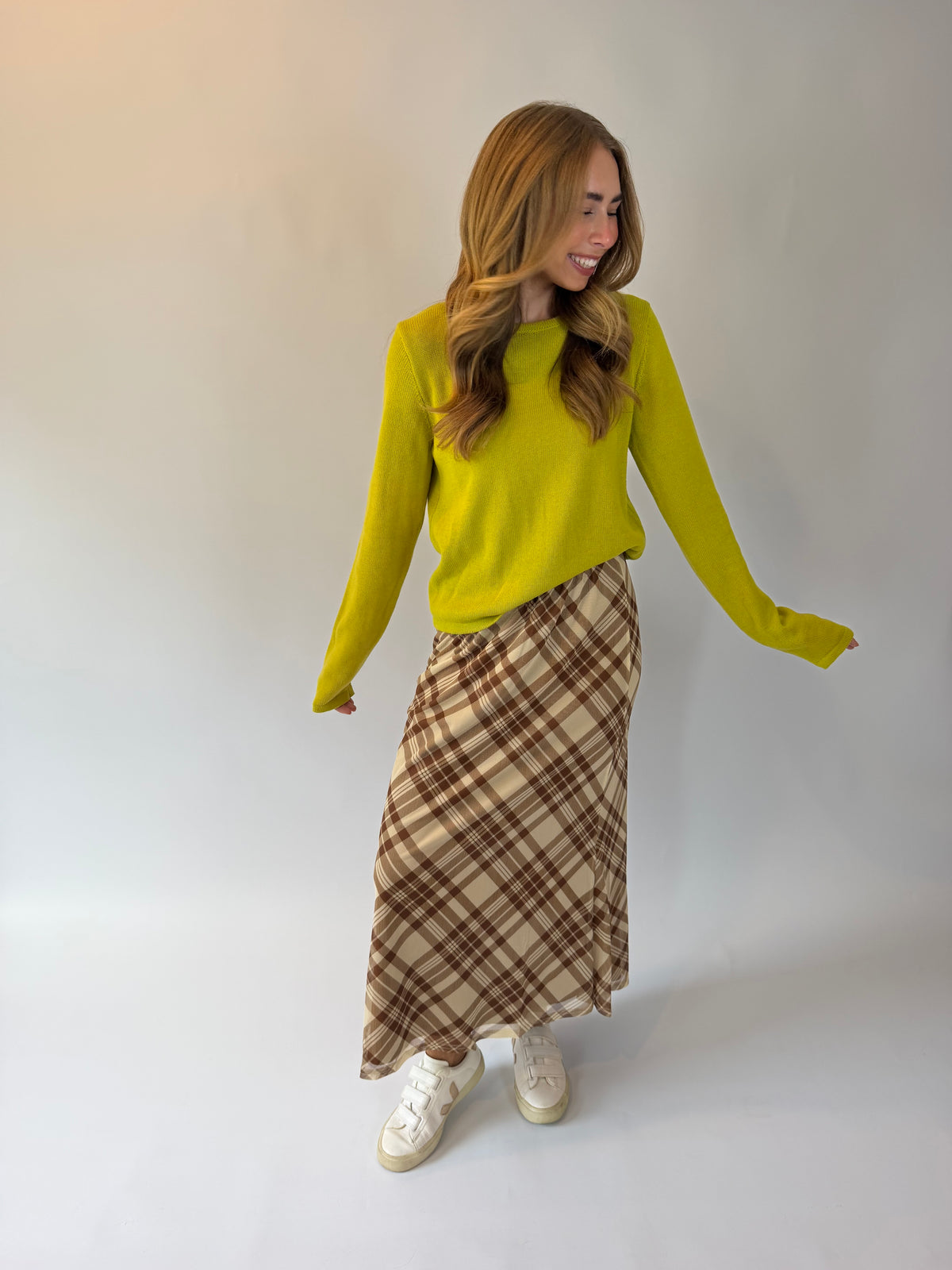 A woman modeling a bright yellow knit sweater and a brown and beige plaid midi skirt with white sneakers, standing against a plain light gray studio background.
