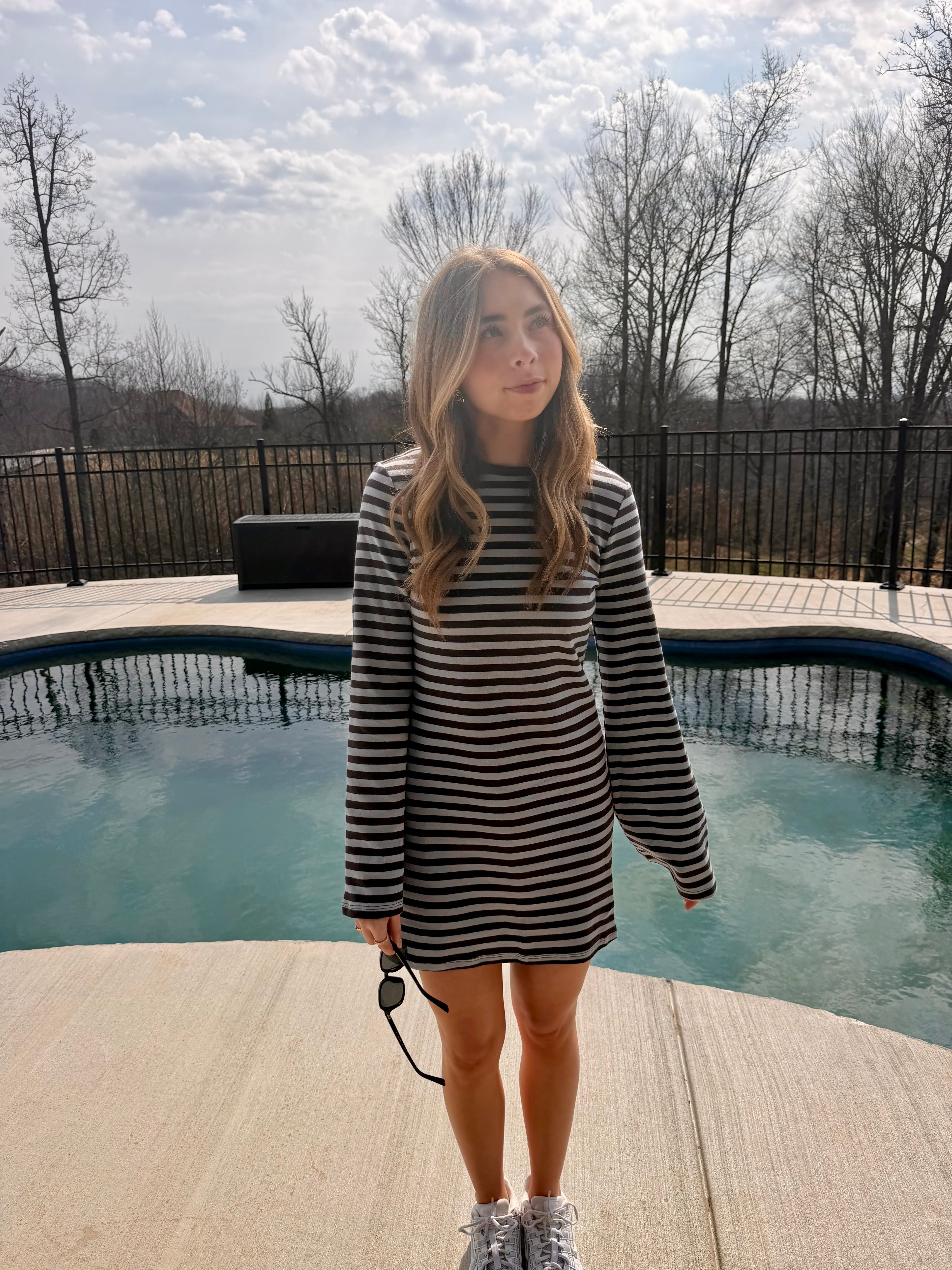 Woman in a striped dress standing by a pool with trees and a cloudy sky in the background