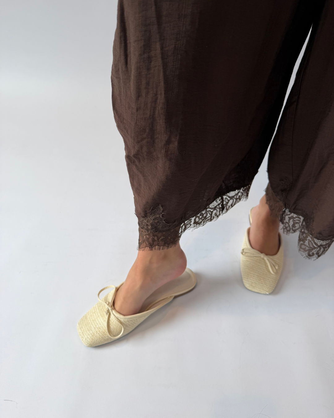 A person wearing loose dark brown draped pants with lace trim and cream woven slip-on mule slippers, shown against a clean light gray studio background.