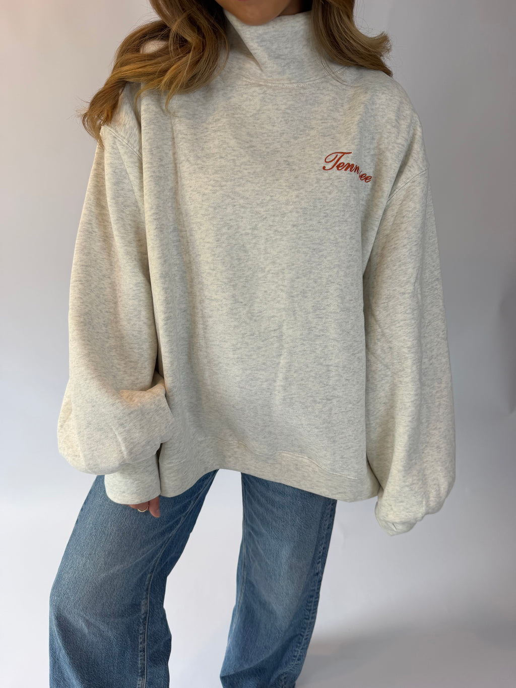 A person wearing an oversized light heather gray turtleneck sweatshirt with red cursive embroidery on the left chest, paired with blue jeans, against a plain light gray background.