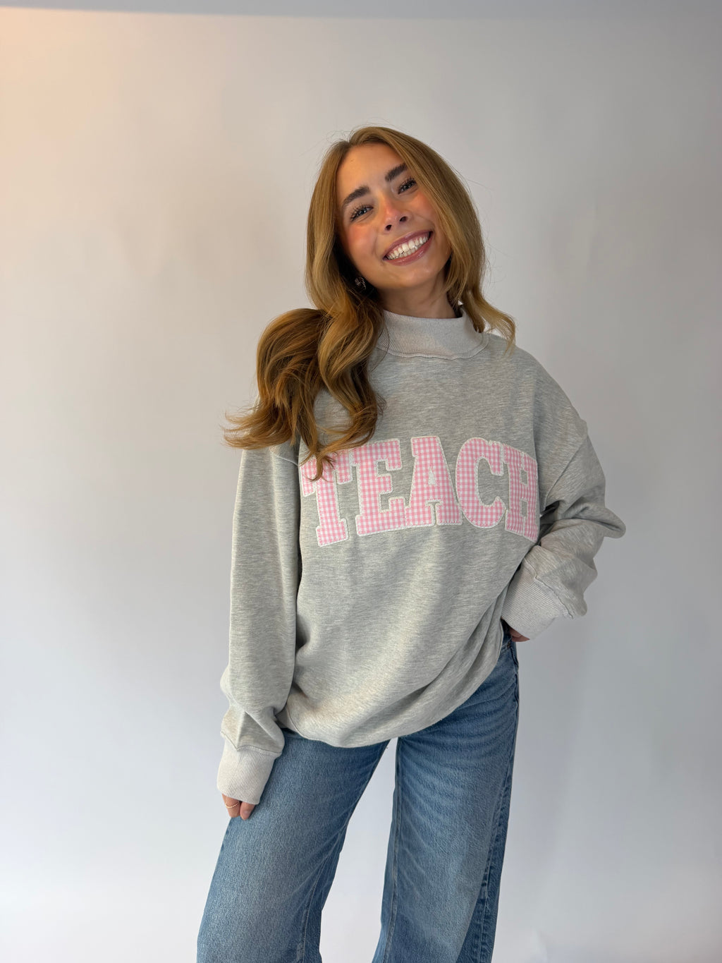 A woman wearing a light gray crewneck sweatshirt with 'TEACH' printed across the chest in pink gingham letters, paired with blue jeans, standing against a plain light gray background.