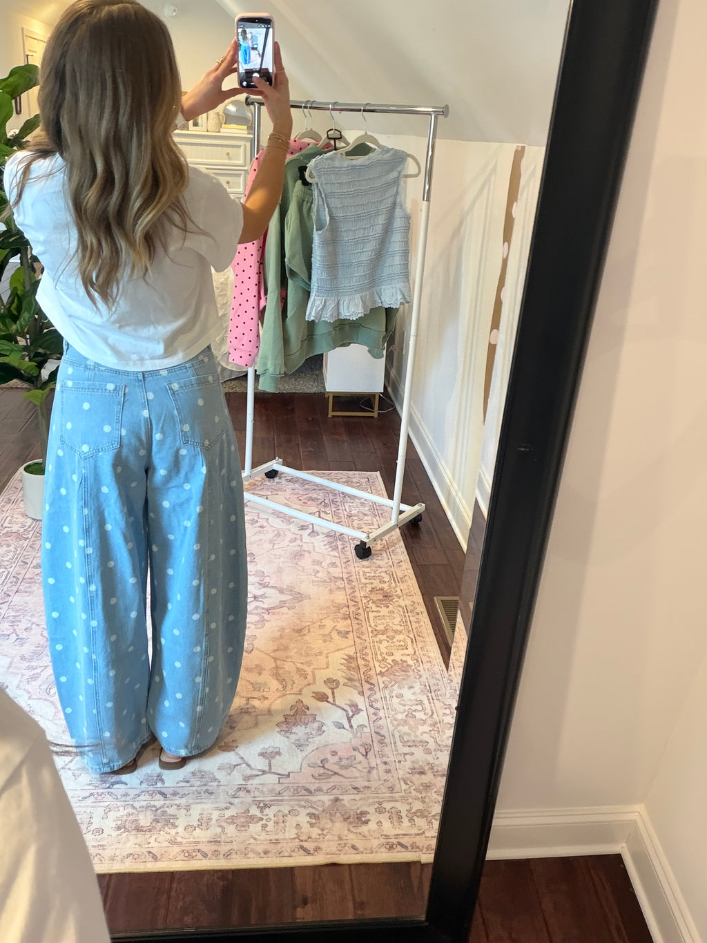 Person taking a mirror selfie wearing light blue pants with white polka dots in a room with a clothing rack.
